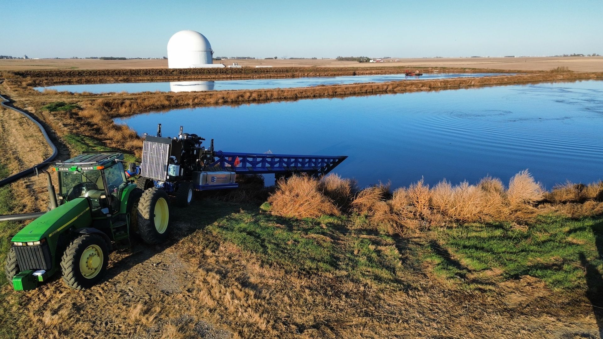 Green tractor spreading material beside a blue pond with a white dome building in the background