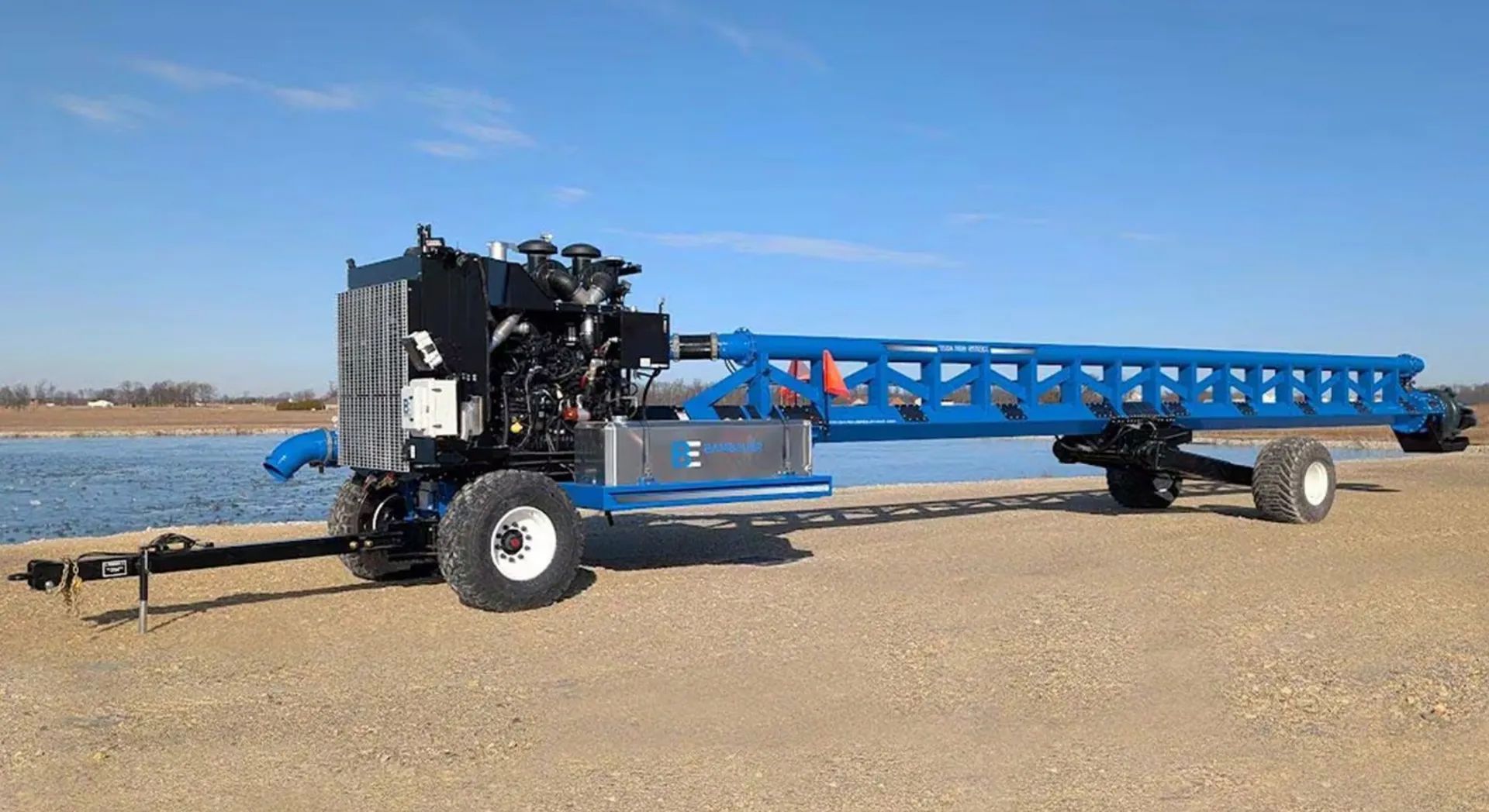 Blue agricultural conveyor trailer on a gravel roadside near water under a clear sky