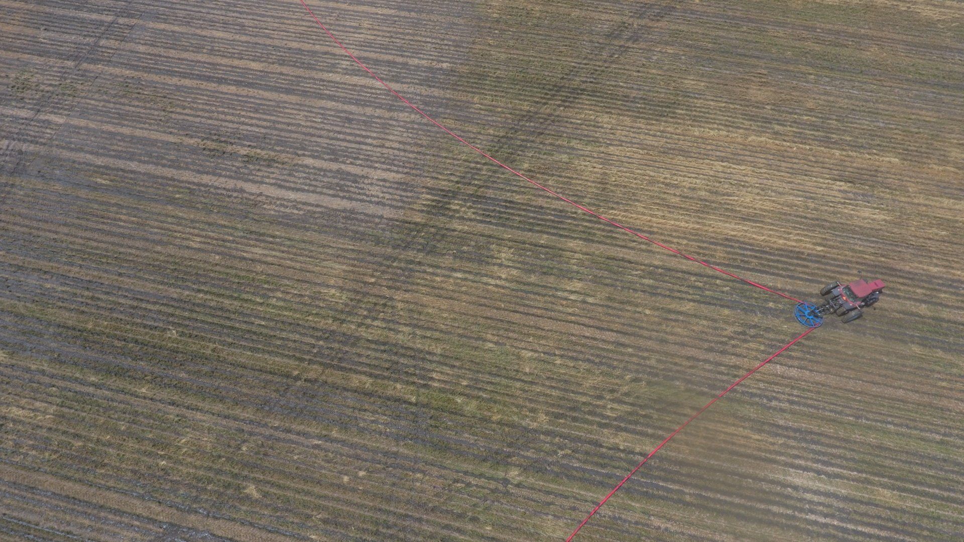 Aerial view of a striped farm field with a small red and blue tractor near the right side.