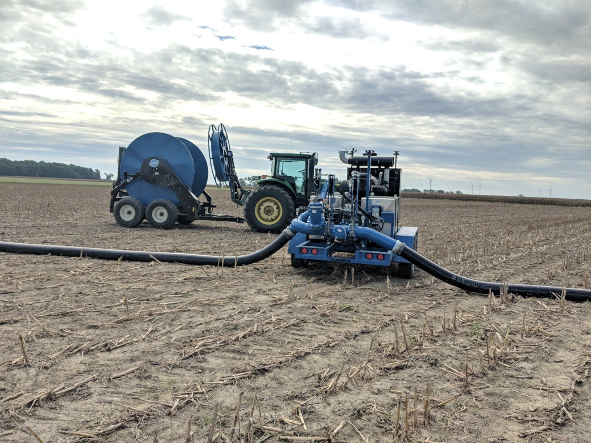 Tractor with blue farm equipment laying irrigation hose across a bare field under cloudy sky