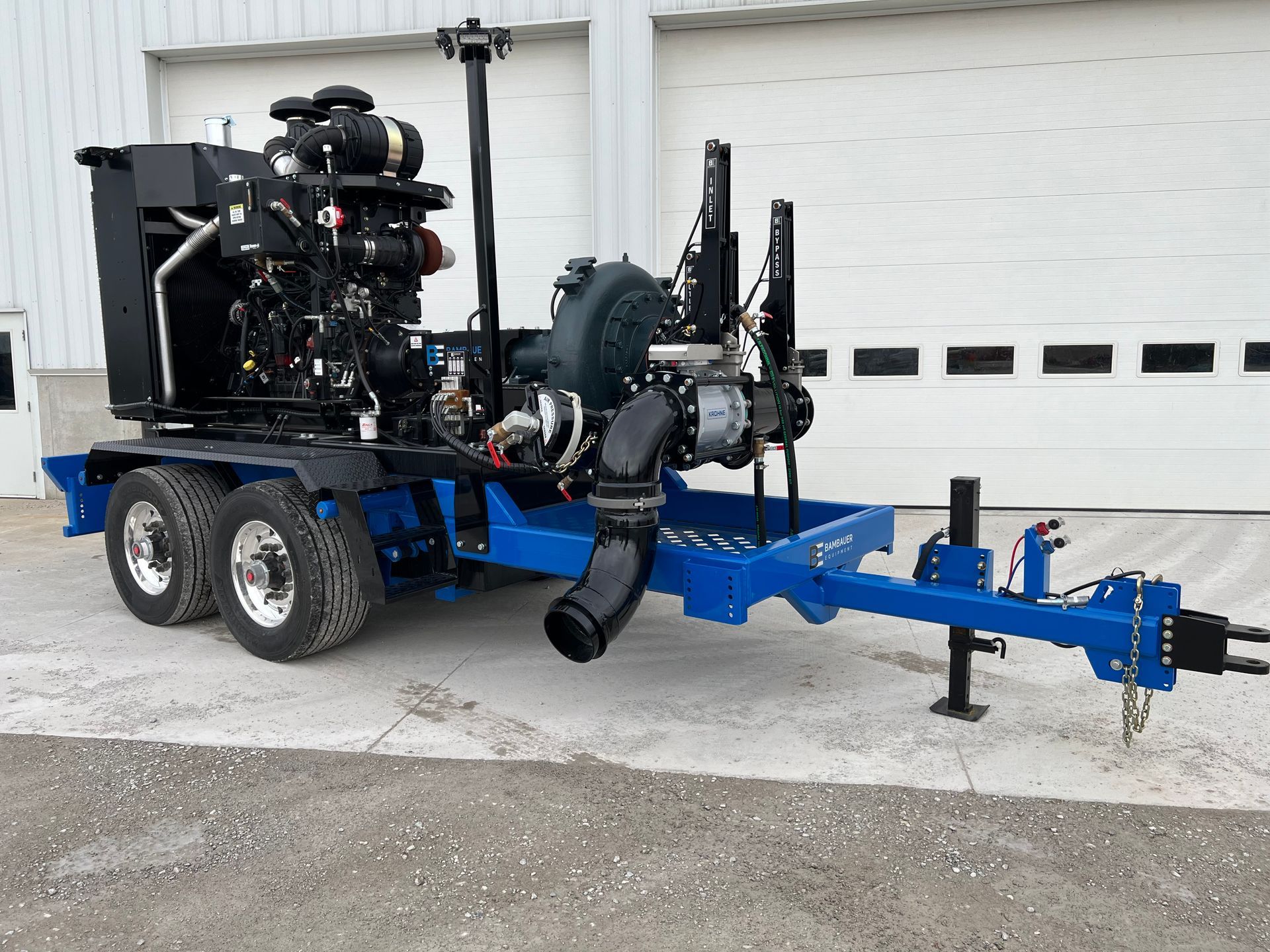 Blue dual-axle trailer carrying a large industrial pump and engine parked beside a white garage door