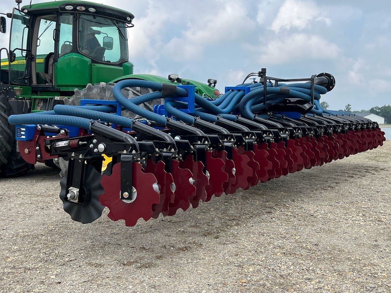 Green tractor pulling a wide red tillage implement with blue hoses on a gravel lot