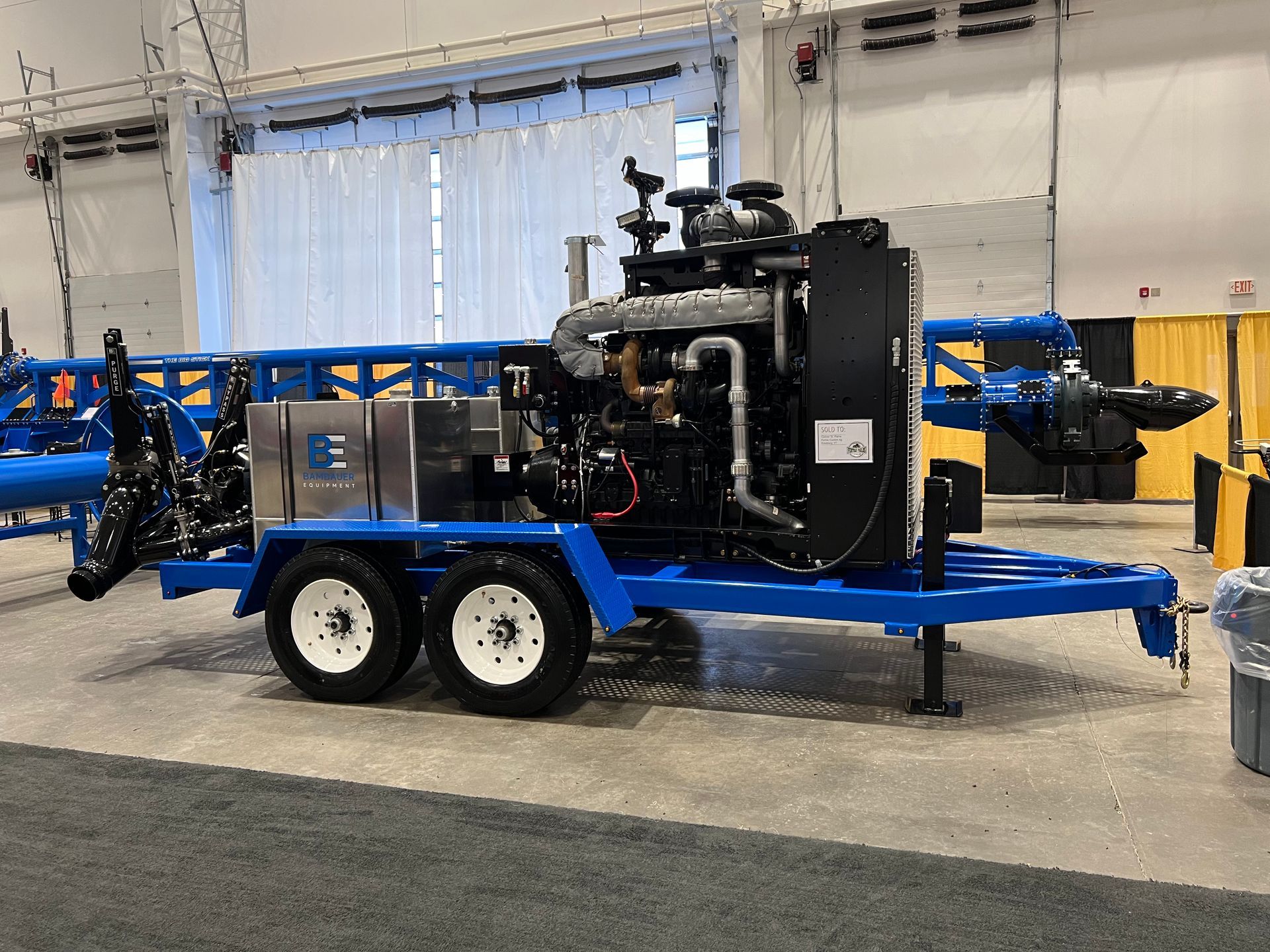 Blue and black industrial machine trailer with dual axles displayed indoors at a trade show.