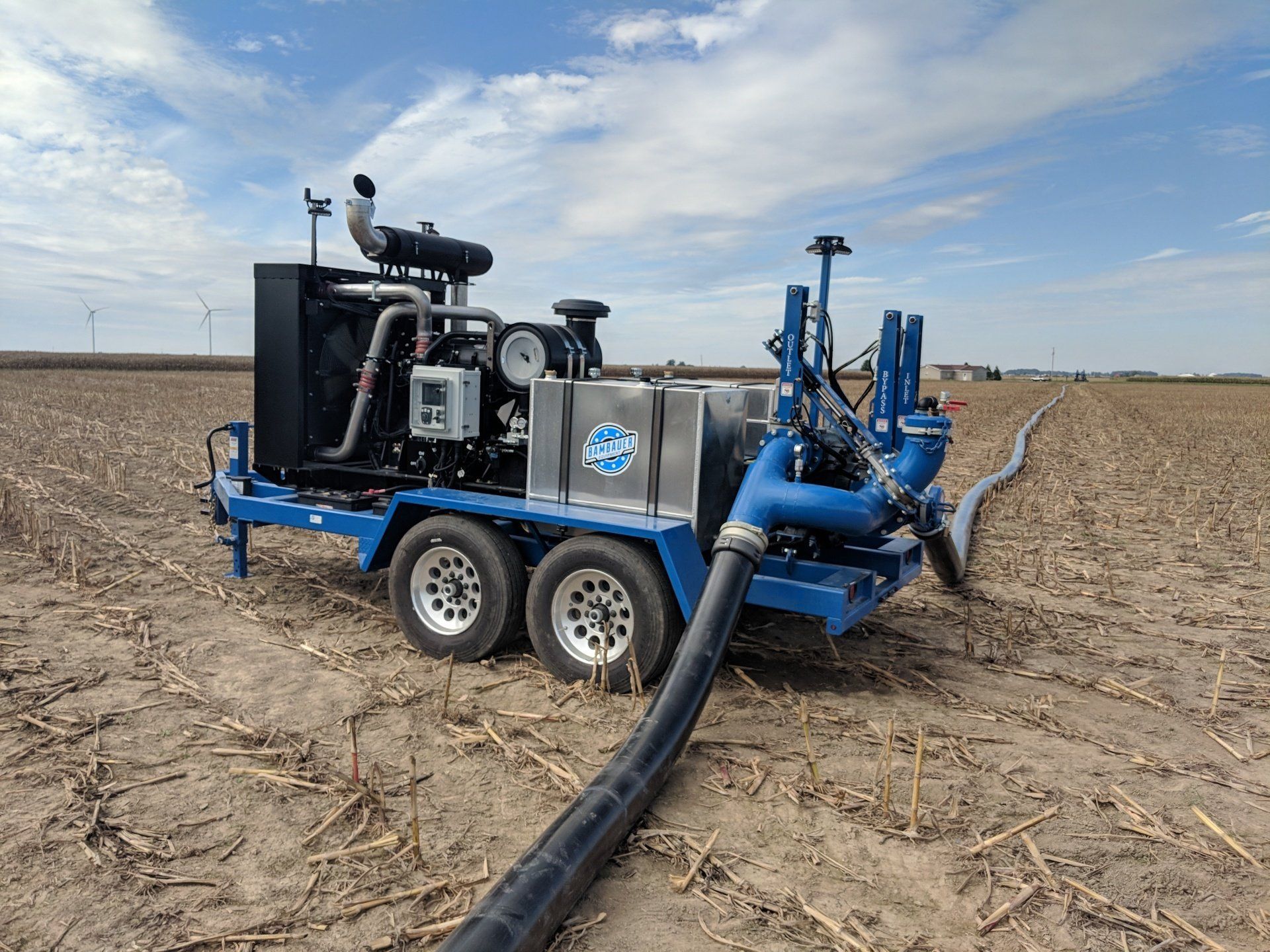 Blue agricultural vacuum truck in a harvested field under a cloudy sky, hose extended to the ground