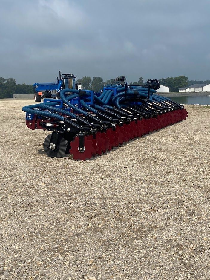 Blue farm implement with red tillage blades parked on gravel under a cloudy sky