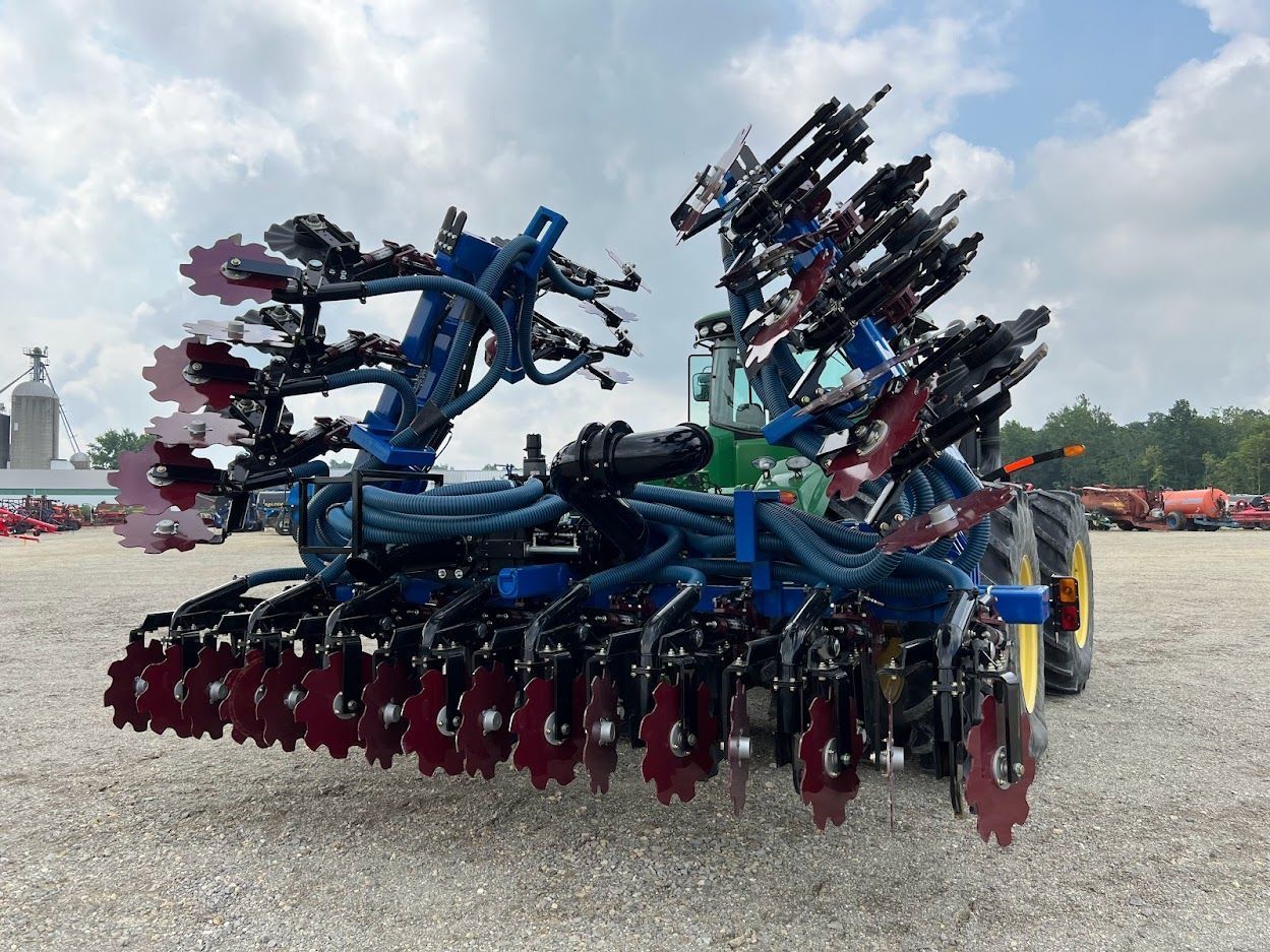 Large blue farm implement with red wheels parked on gravel under a cloudy sky