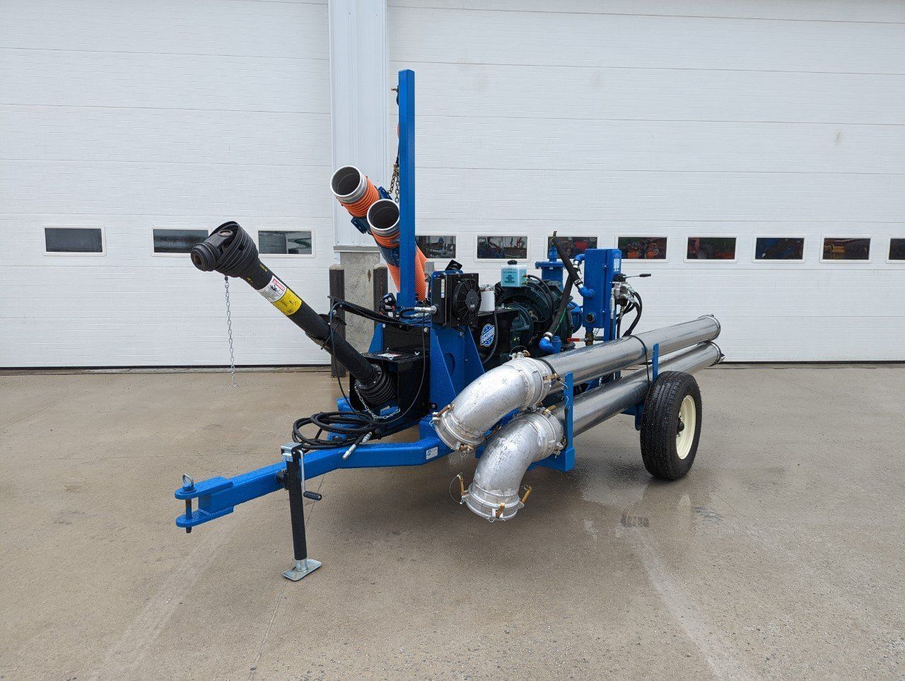 Blue agricultural machine with large silver pipes and wheels parked on concrete in front of a garage door