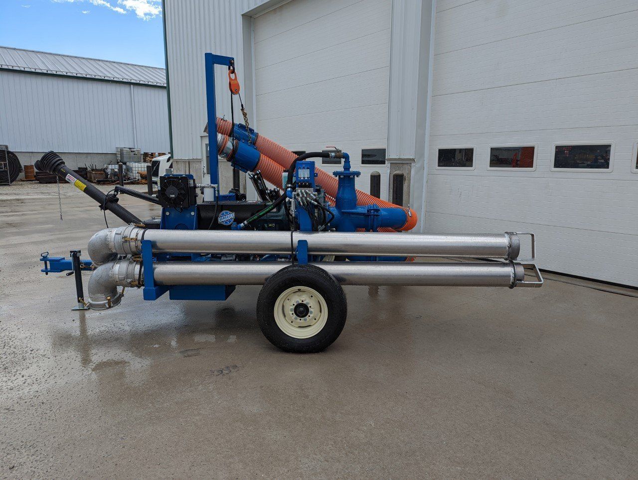 Blue industrial trailer with metal pipes and equipment parked outside a warehouse on wet pavement