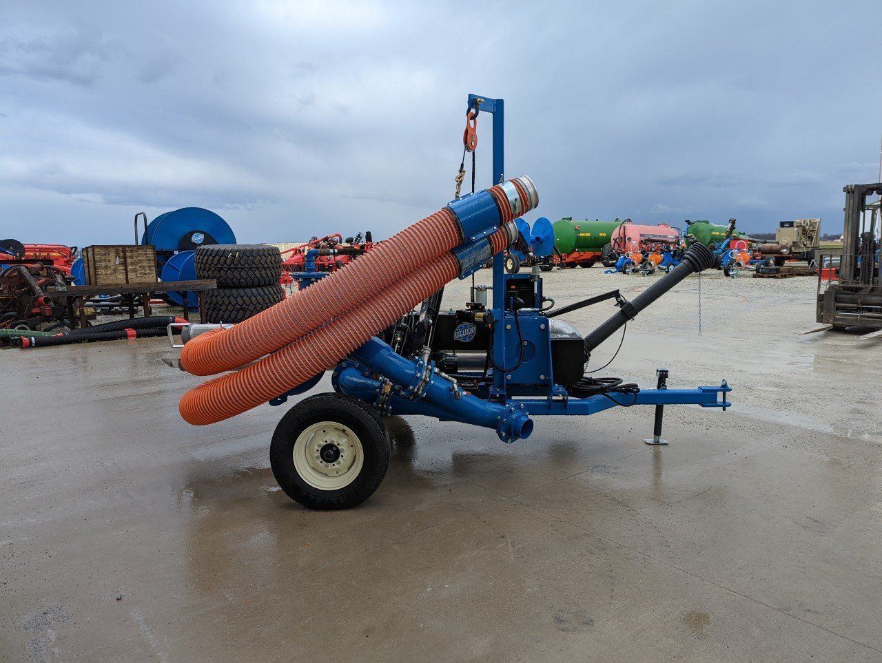 Blue agricultural machine with orange flexible tubes parked on a wet lot under cloudy skies