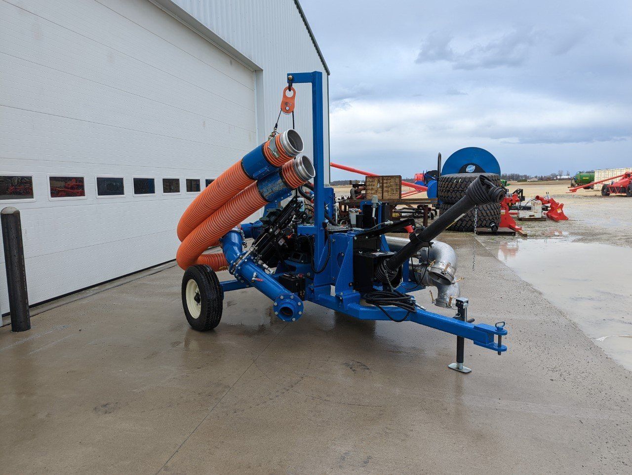 Blue agricultural machine with orange hoses parked beside a building on a wet outdoor lot