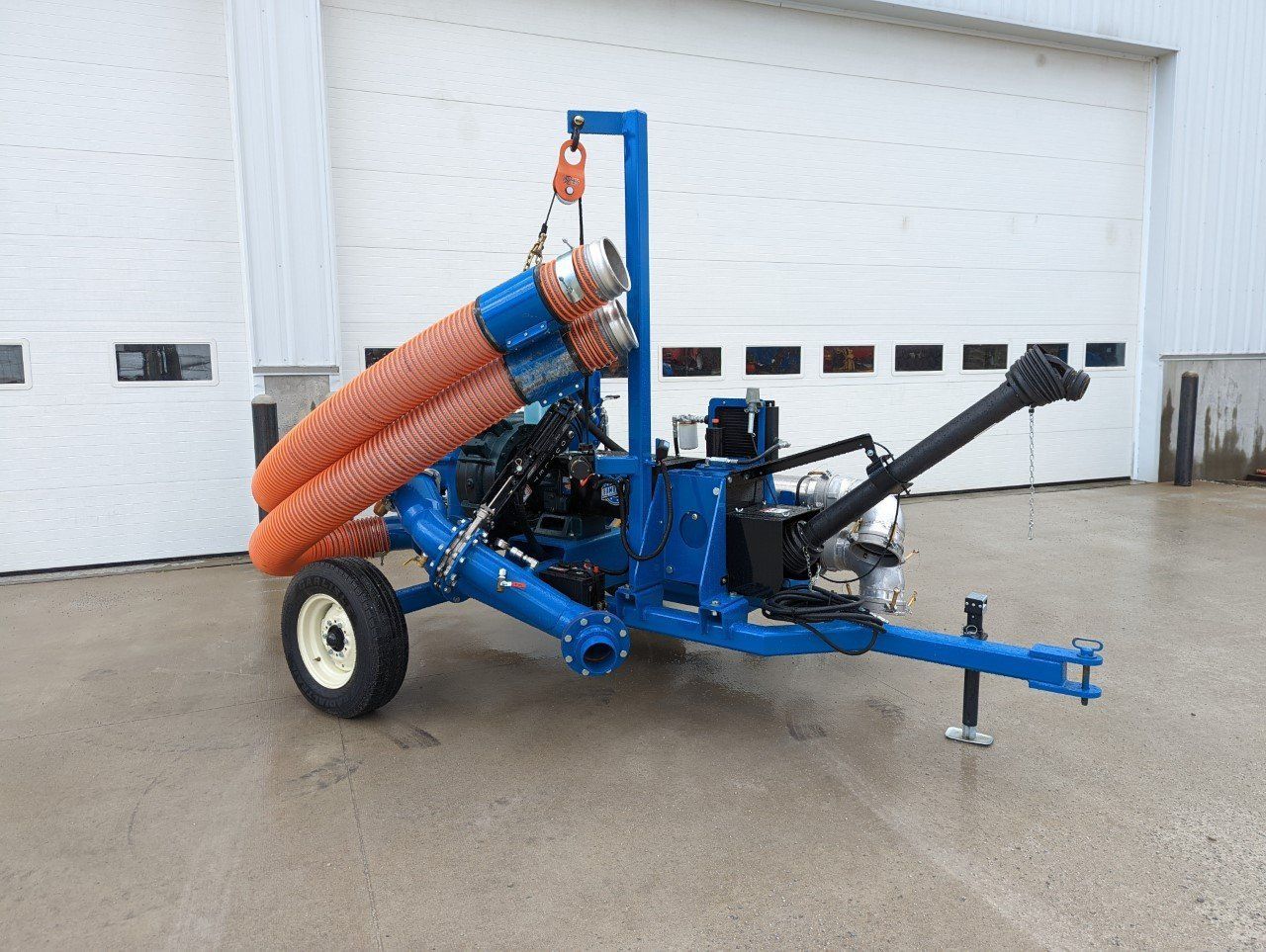 Blue agricultural trailer with orange hoses parked on wet pavement outside a white garage door