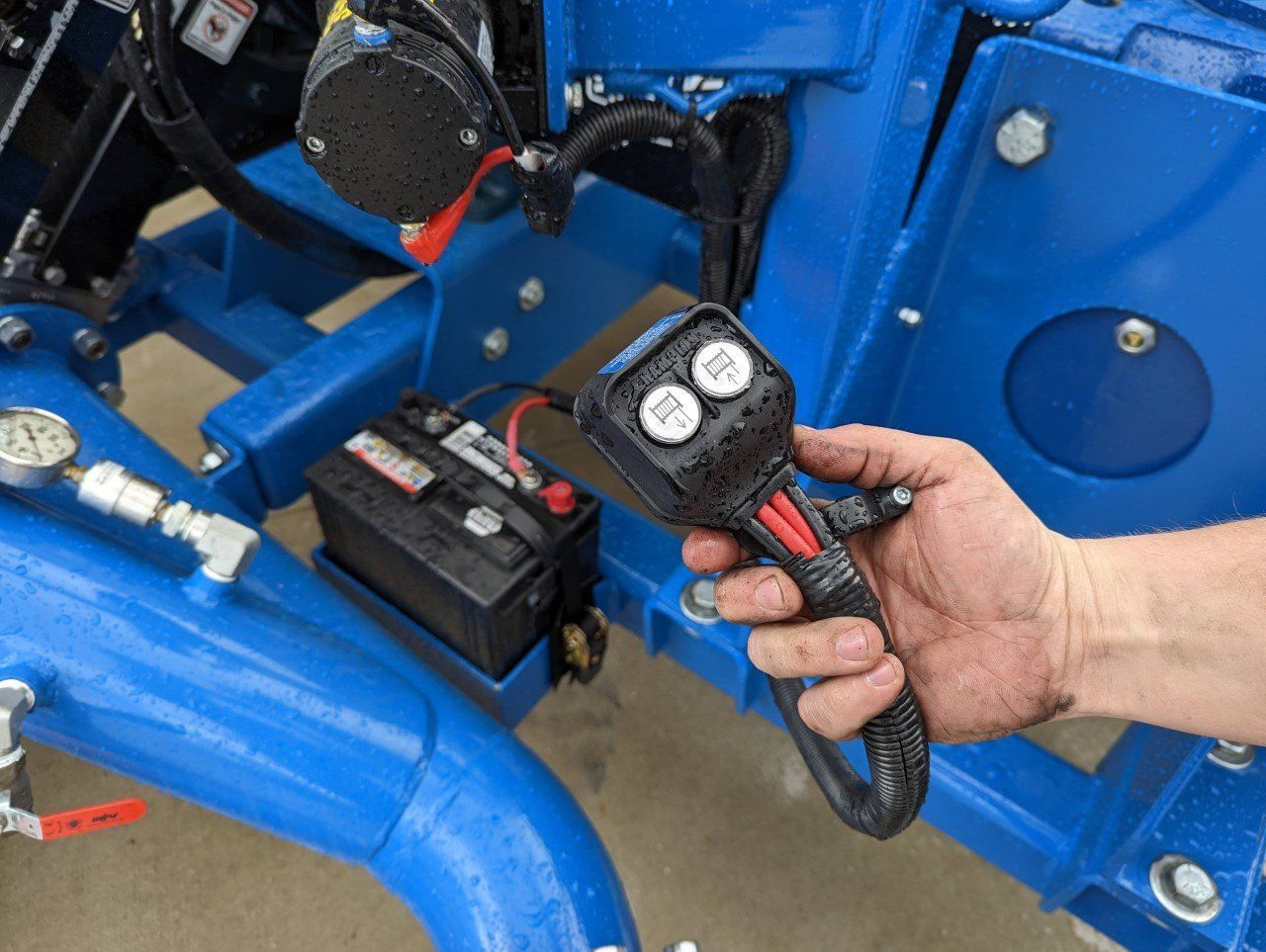 Hand holding a wired dual-gauge remote control near blue machinery and a battery pack