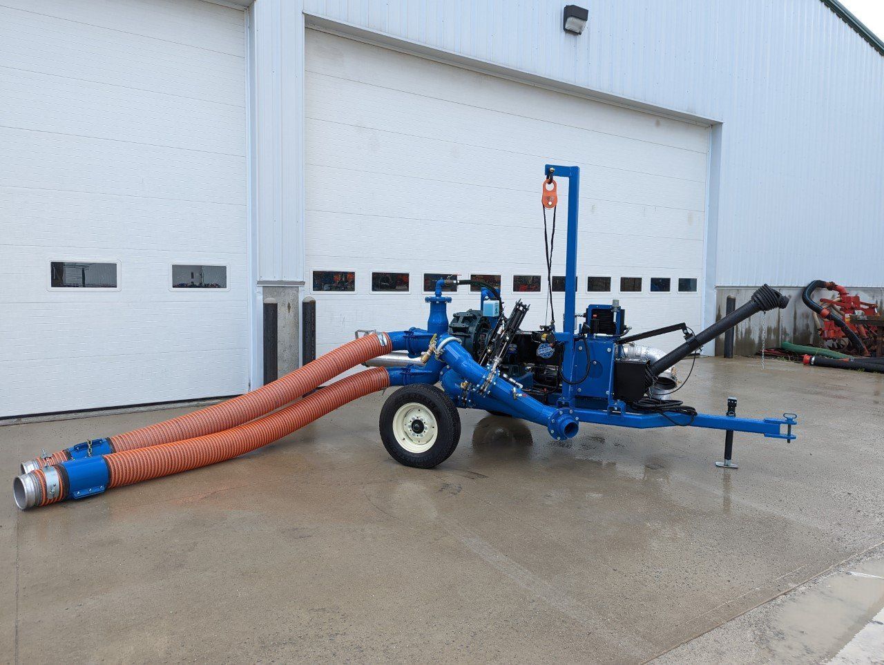 Blue industrial vacuum trailer with orange hoses parked outside a white building on wet pavement