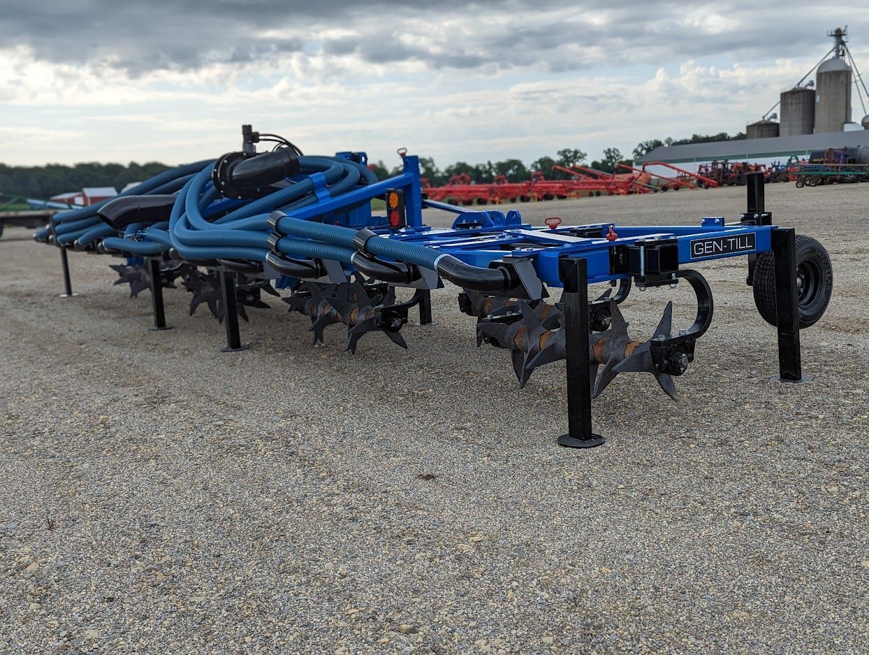 Blue agricultural tillage implement with black tines parked on gravel outdoors