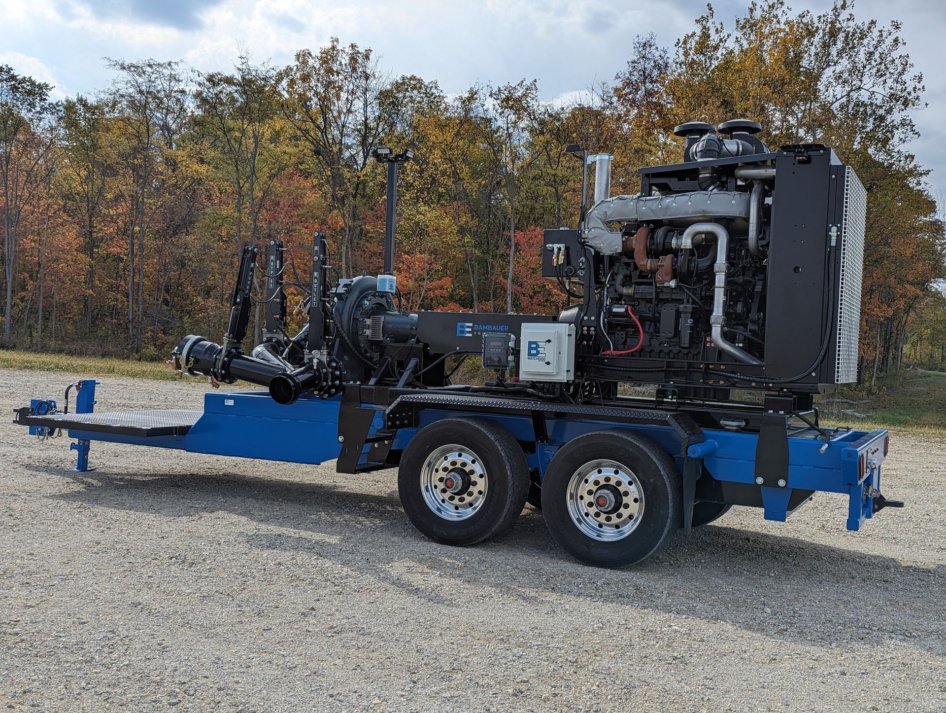 Blue trailer-mounted industrial machine with engine and wheels parked on gravel in front of autumn trees