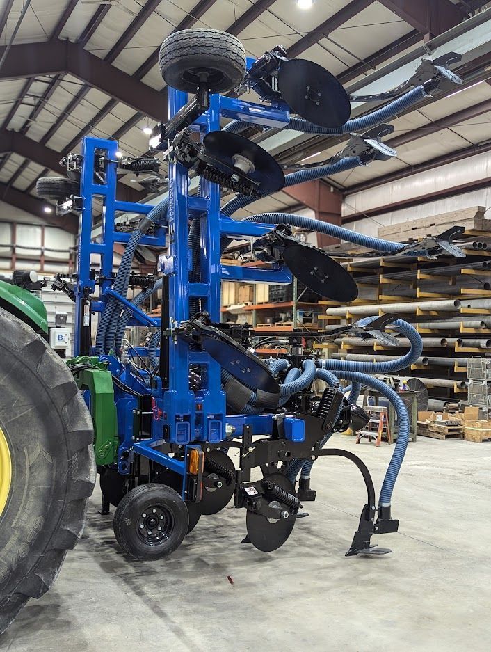 Blue farm implement with large black discs and wheels inside a warehouse, beside a tractor tire