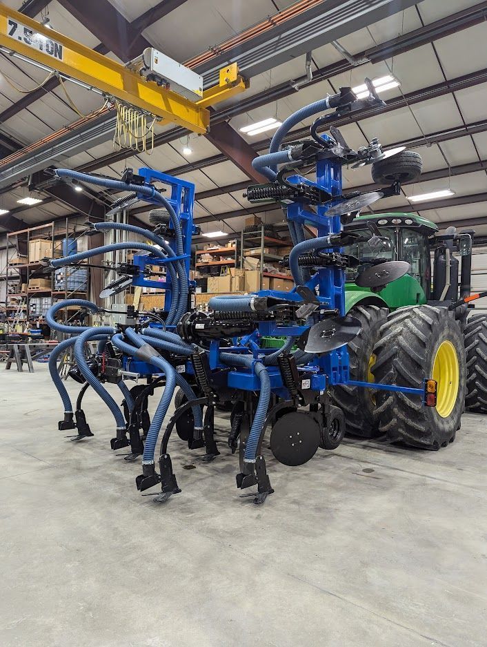 Blue agricultural machine with folded arms inside a warehouse, attached to a green tractor