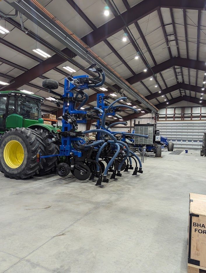 Blue agricultural machine with large black hoses in a warehouse, beside a green tractor.