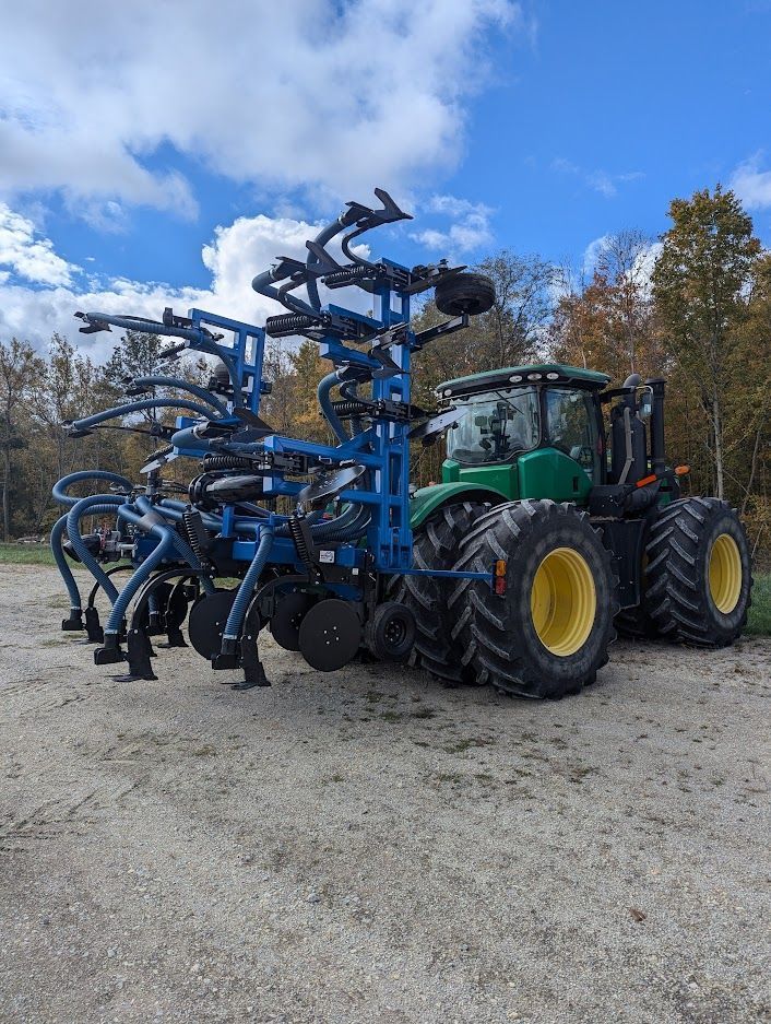 Green tractor pulling a large blue farming implement on a gravel road under a cloudy sky