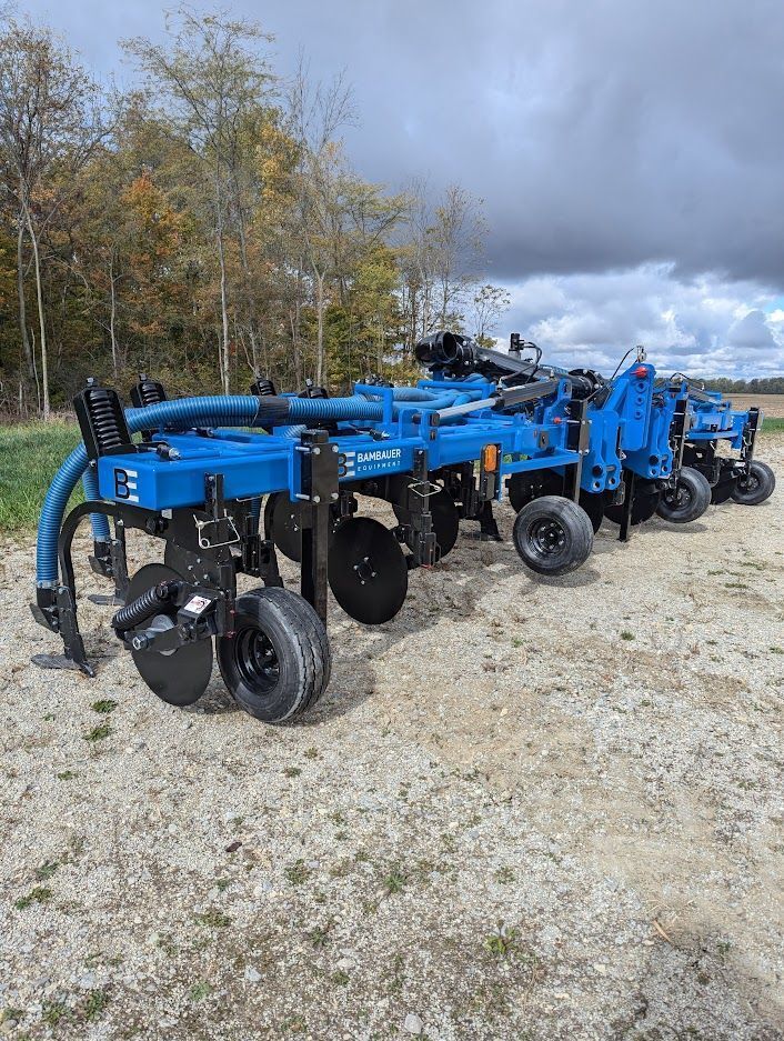 Blue agricultural tillage equipment lined up on a gravel lot near trees under a cloudy sky