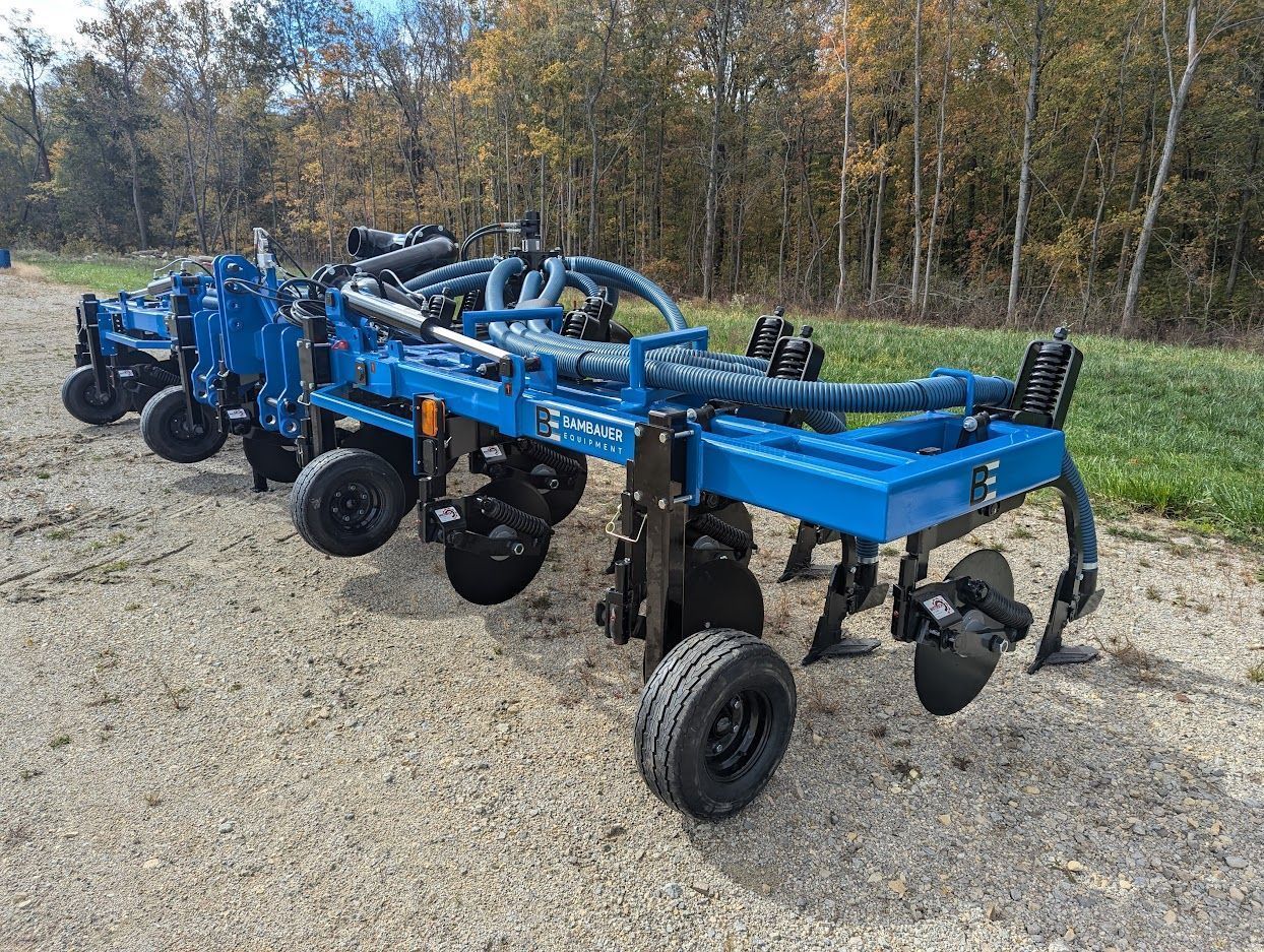 Blue agricultural tillage implement on wheels parked on gravel with trees in the background