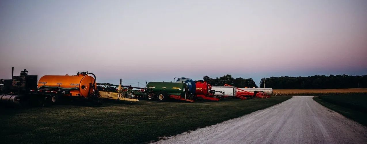 Long line of trucks and trailers parked beside a gravel road at dusk under a pink sky