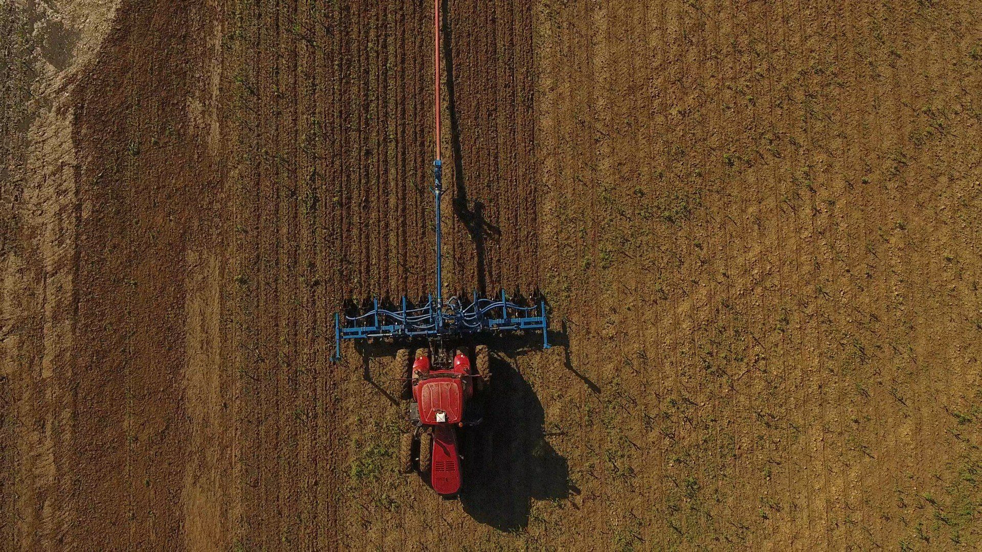 Aerial view of a red tractor plowing a brown field, leaving a narrow trail behind it.