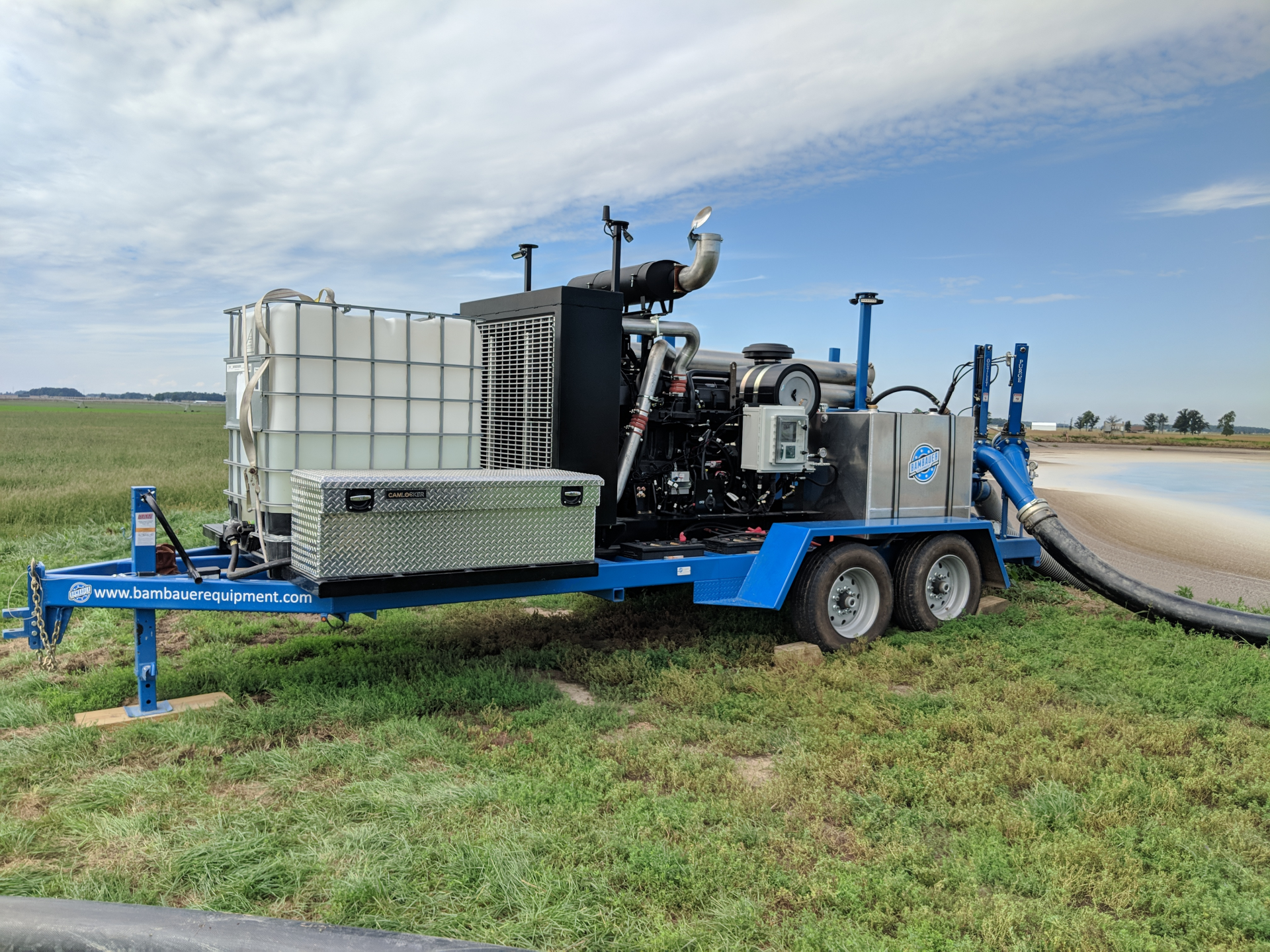 Blue pump truck with large tank and hose on a grassy field under a cloudy sky