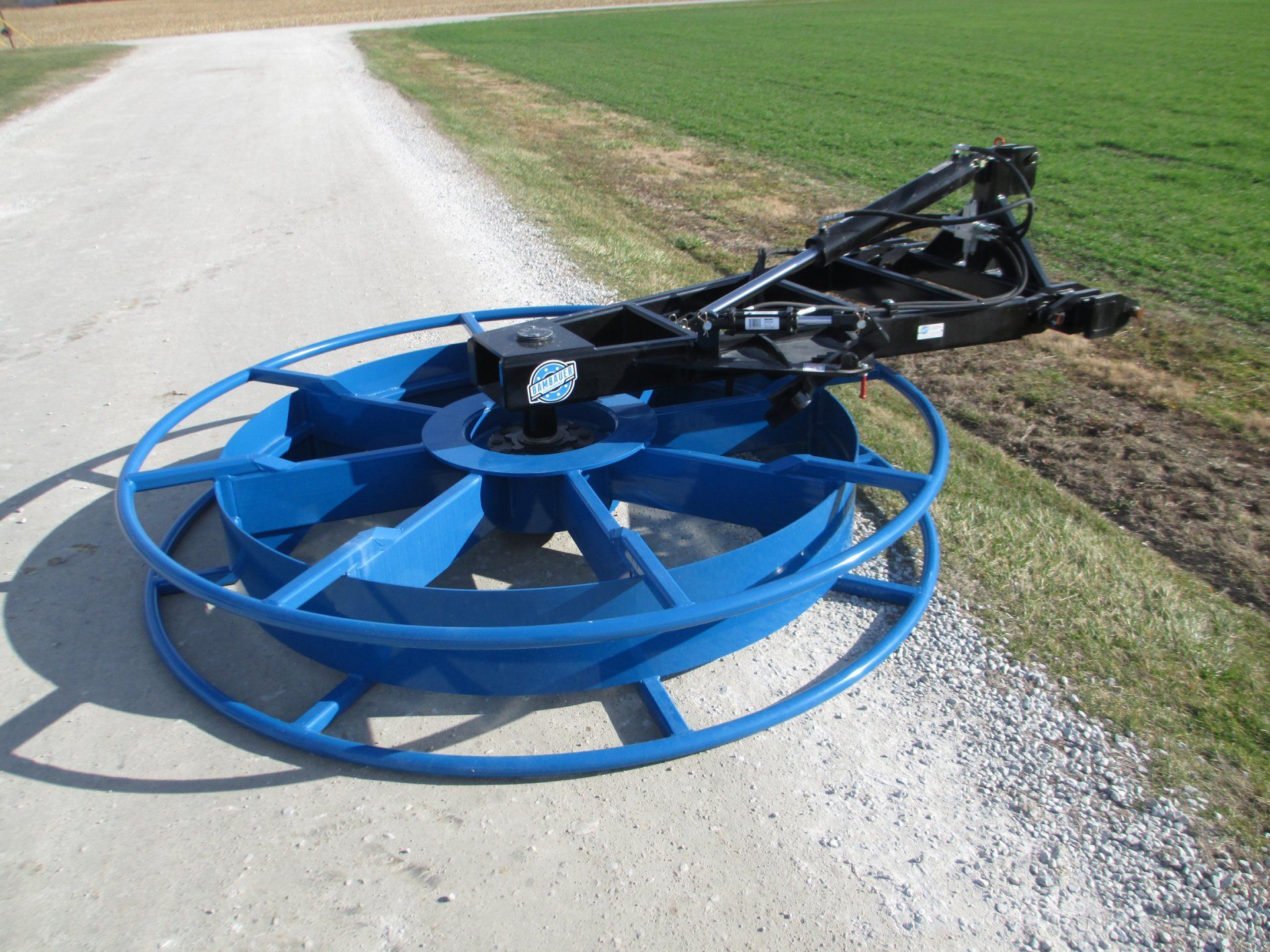 Blue rotary mower attachment on a gravel roadside beside grass fields.