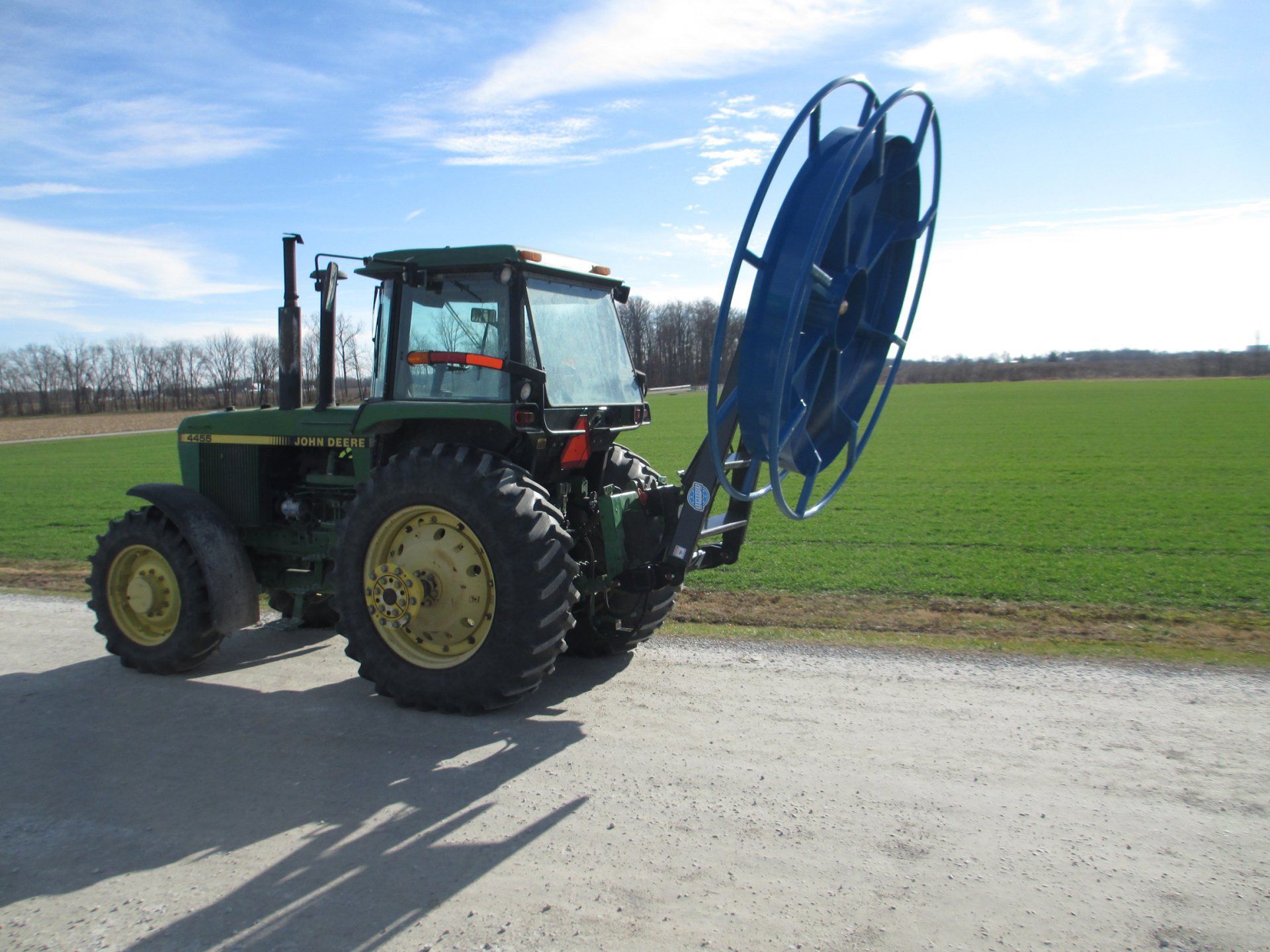 Green tractor with large blue hose reel on a rural road beside a field under a clear sky
