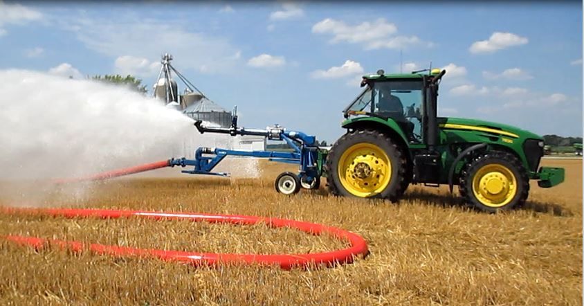 Green tractor spraying a field with water from a boom sprayer under a blue sky.
