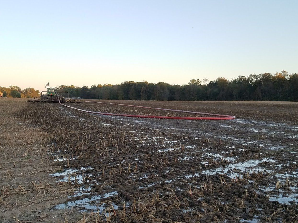 Plowed field with patches of snow at sunrise, farm machinery in the distance, and a red hose across the ground