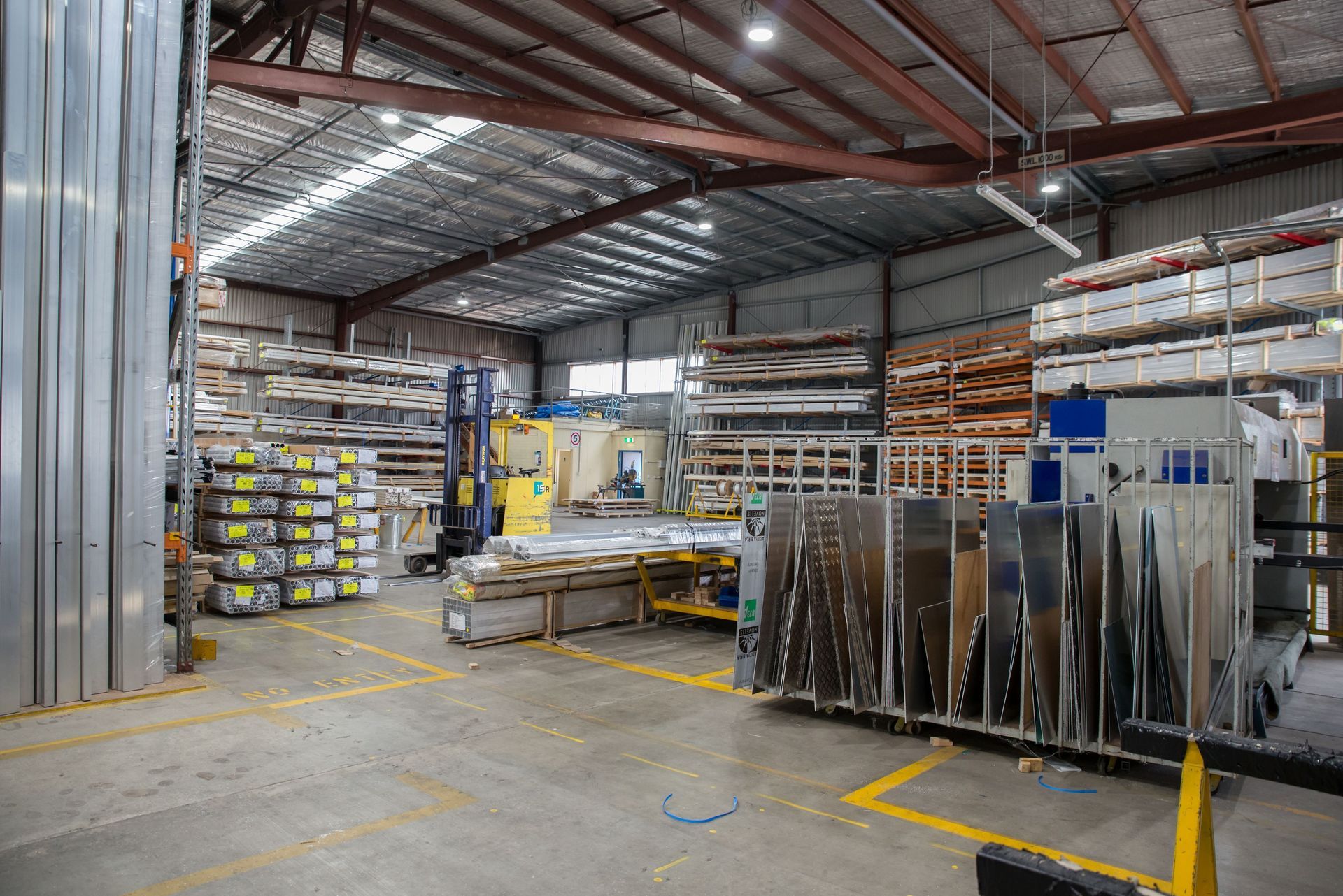 Industrial warehouse interior with stacked metal sheets, shelving, and yellow safety markings on the floor