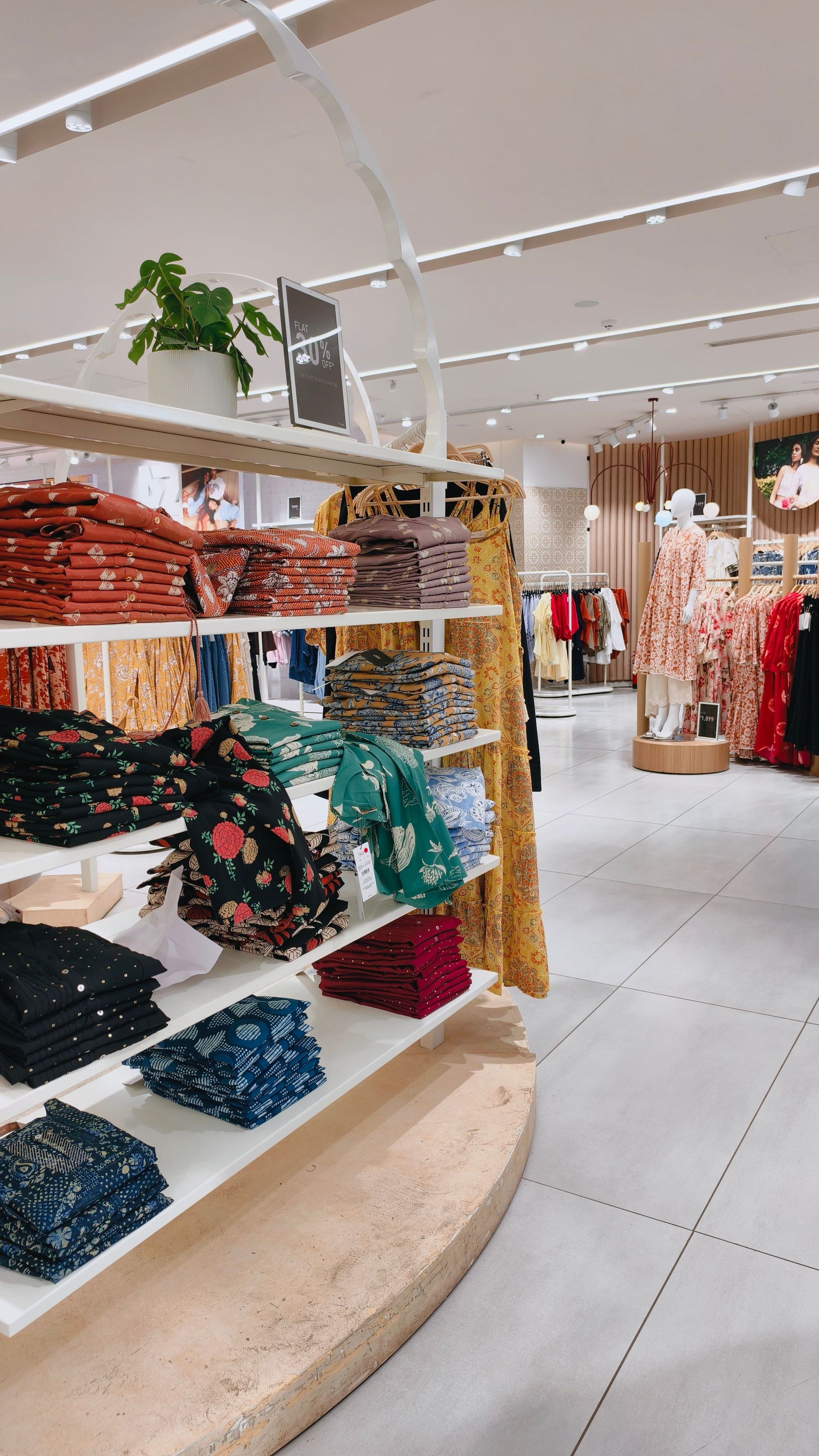 Bright clothing store aisle with folded patterned sweaters on shelves and hanging dresses in the background