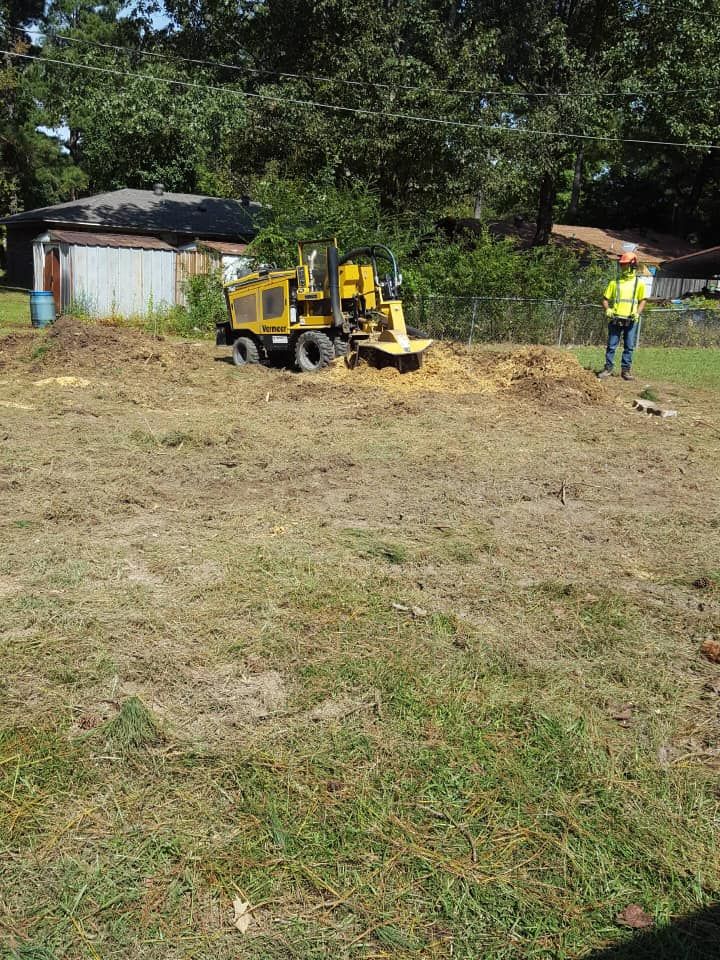 A yellow stump grinder sits on a dirt mound in a grassy yard, with a person in high-visibility gear standing nearby.