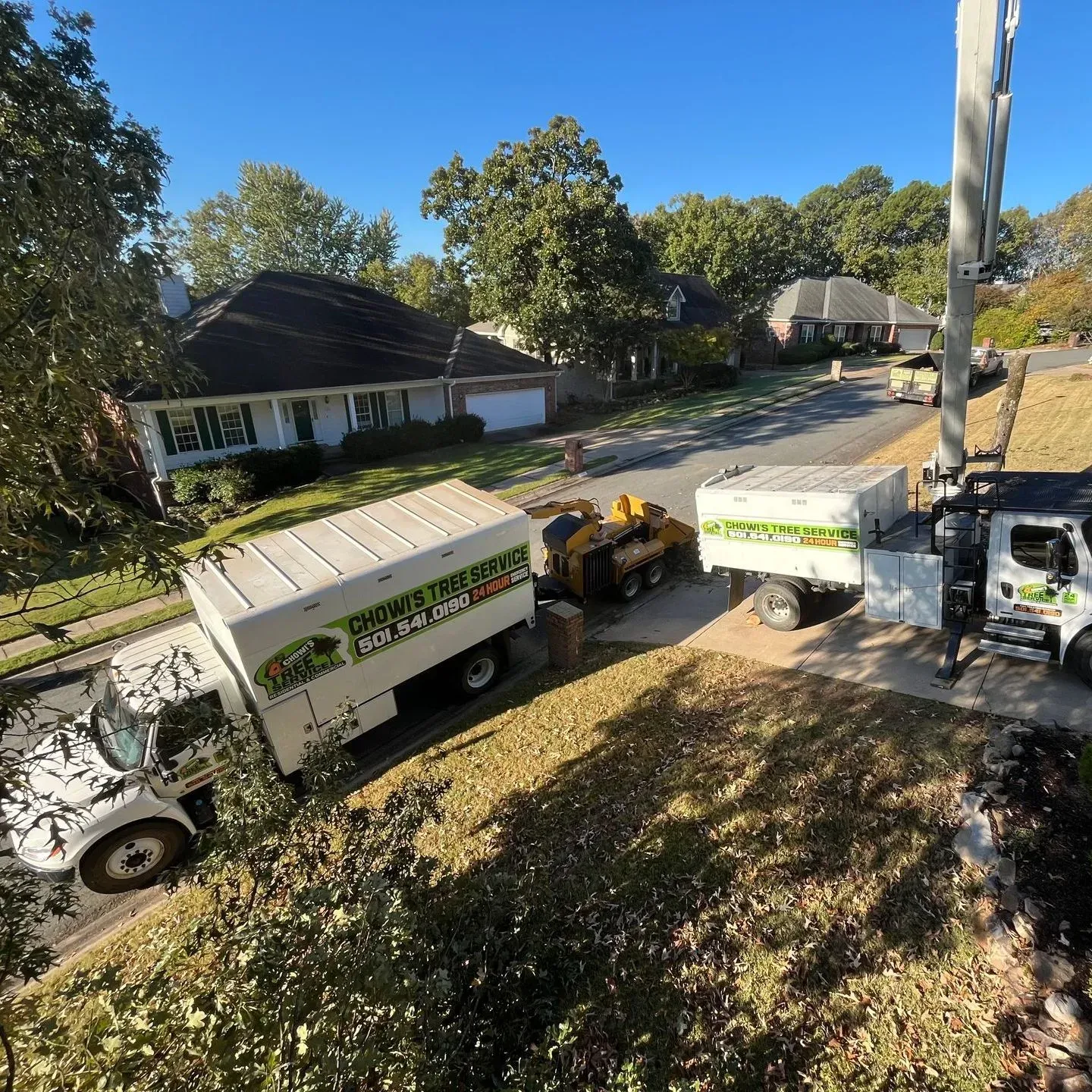 A work crew with white trucks and equipment is trimming trees on a residential street in a suburban neighborhood.