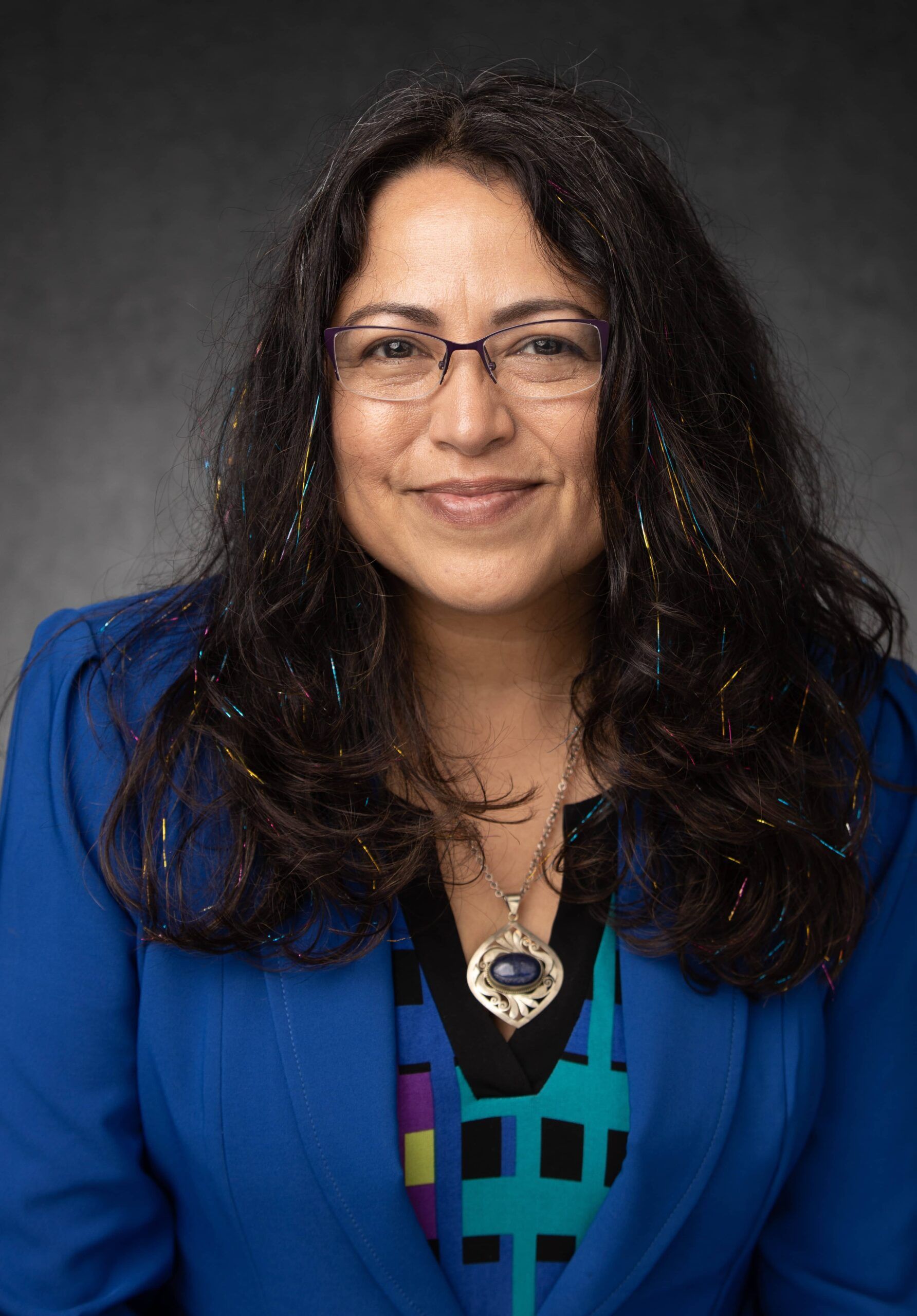 A woman with long, wavy black hair wearing a vibrant blue blazer, colorful patterned blouse, and a silver necklace with a blue stone pendant. She has glasses and a warm smile, with subtle, colorful tinsel strands in her hair. The background is a plain dark gray.