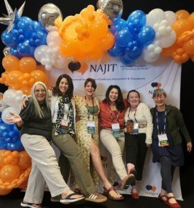 Five women posing at a conference booth with orange, blue, and white balloon decorations and a NAJIT banner