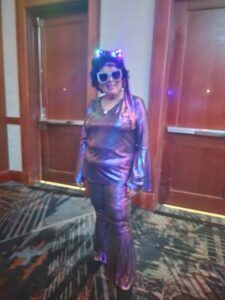 Person in a shiny metallic outfit and sunglasses posing in front of wooden double doors.
