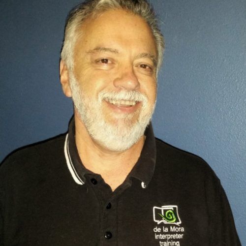 Smiling man with gray beard in a black polo shirt against a blue wall