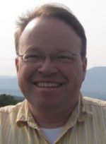 Smiling person in glasses and a striped shirt outdoors with mountains in the background