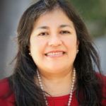 Smiling person with long dark hair wearing a red top and pearl necklace outdoors