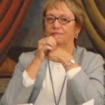 Woman with glasses resting her chin on her hands, wearing a light blue top and lanyard, seated before a painted backdrop