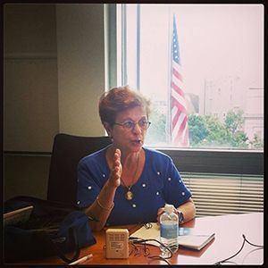Woman speaking at a conference table in a meeting room, with a window and American flag behind her.