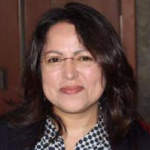 Portrait of a smiling person with dark hair, glasses, and a black-and-white patterned blouse.
