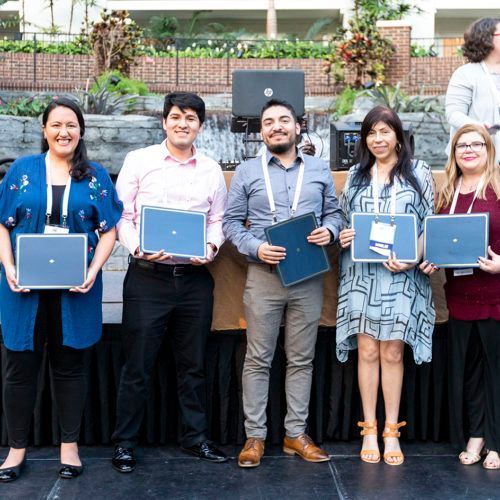 Five adults holding laptops and tablets, standing together outdoors in front of a building