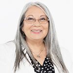 Smiling person with long silver hair and glasses, wearing a black-and-white polka dot top on a white background