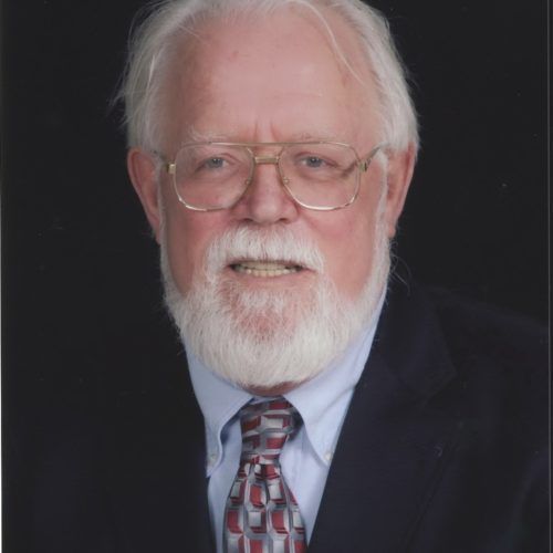 Portrait of a white-bearded man in glasses, suit, and patterned tie against a dark background