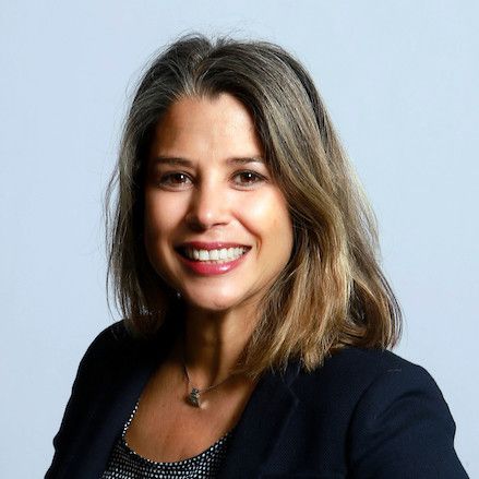Smiling woman with shoulder-length brown hair in a dark blazer against a light blue background