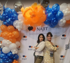 Two women pose back-to-back before a NAJIT banner and blue, orange, and white balloons.
